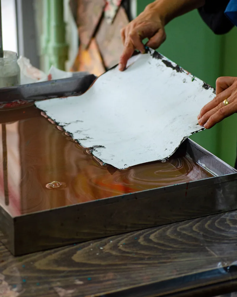 Hoja de papel todavía húmeda levantada con las dos manos sobre el borde de una tina de taller